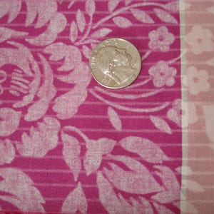 May include: Close-up of a pink fabric with a floral pattern and a coin. The fabric features large white floral designs and smaller flowers. The coin is silver and shows a profile.