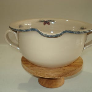 May include: Cream ceramic bowl with two handles and a scalloped rim with blue trim. The bowl has a pinecone design and sits on a small, round wooden stand. A decorative kitchen item.