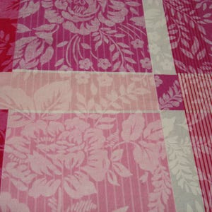 May include: Close-up of a floral fabric with a patchwork design. The pattern features large, stylized roses and foliage in shades of pink and white. The fabric is divided into rectangular sections with varying patterns.