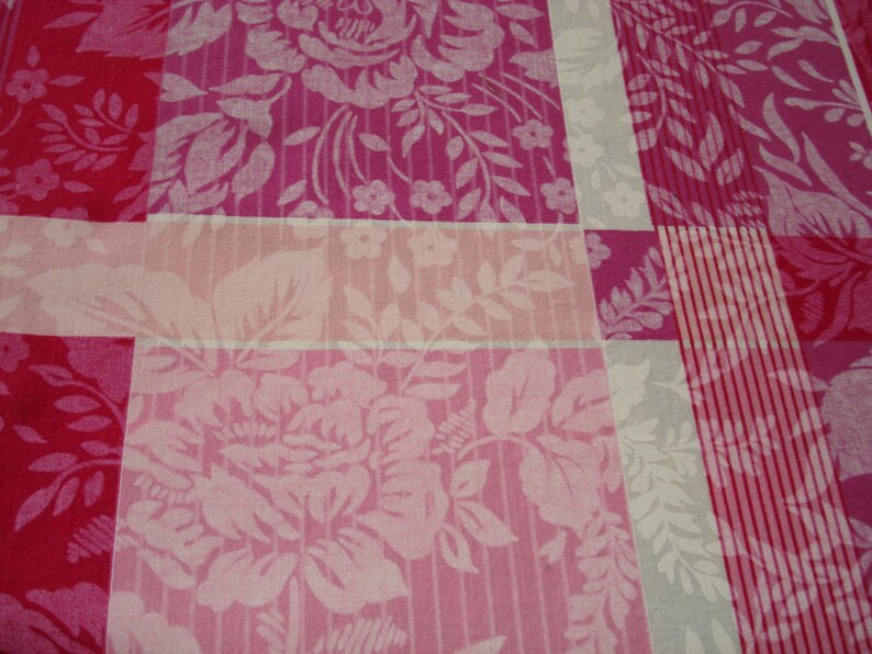 May include: Close-up of a floral fabric with a patchwork design. The pattern features large, stylized roses and foliage in shades of pink and white. The fabric is divided into rectangular sections with varying patterns.