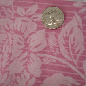 May include: Close-up of pink fabric with a floral design. A silver coin is placed on the fabric for scale. The fabric has a textured appearance with a repeating design of large flowers and leaves.