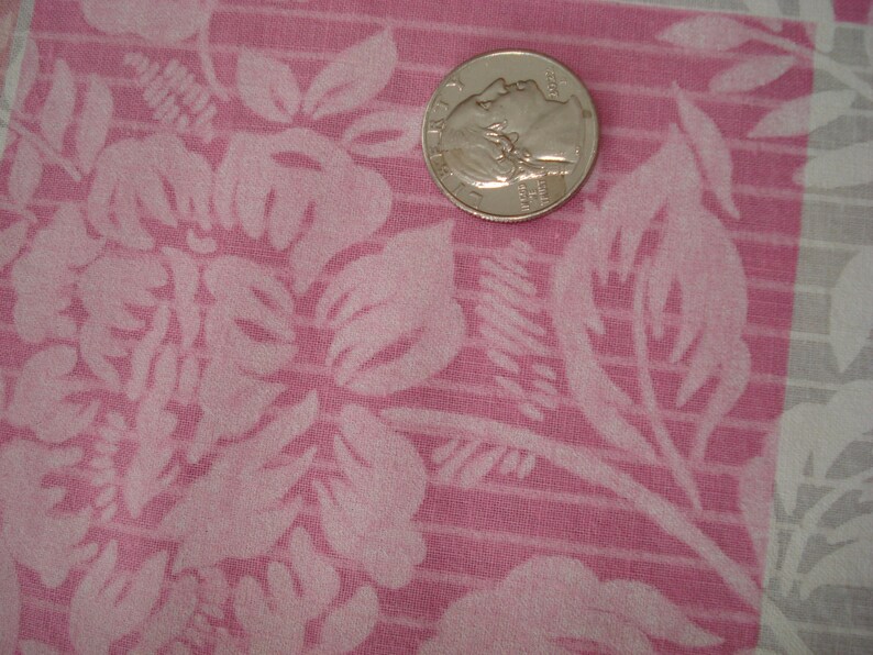 May include: Close-up of pink fabric with a floral design. A silver coin is placed on the fabric for scale. The fabric has a textured appearance with a repeating design of large flowers and leaves.