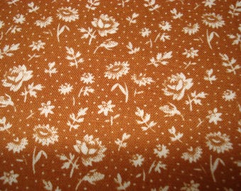 May include: Close-up of a fabric with a repeating floral pattern. The fabric has a warm, orange-brown background with a small, dotted texture. White floral and leaf designs are scattered across the surface, creating a vintage aesthetic.