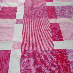 May include: A patchwork quilt in shades of pink. The design features rectangular blocks of varying pink tones, including floral and striped patterns. White strips divide the blocks, creating a visually appealing pattern.