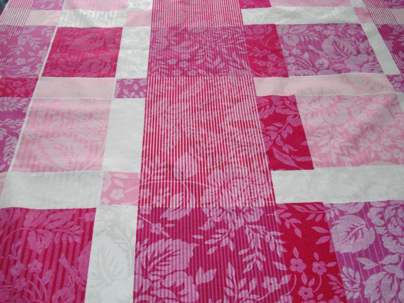 May include: A patchwork quilt in shades of pink. The design features rectangular blocks of varying pink tones, including floral and striped patterns. White strips divide the blocks, creating a visually appealing pattern.