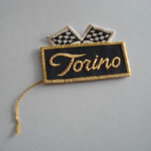 May include: A black and gold embroidered patch with the word "Torino" in gold lettering. The patch has a white and black checkered flag design on top.