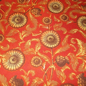 May include: Close-up of a fabric with a repeating floral pattern. The design features large, stylized flowers in shades of brown and gold against a vibrant red background. The fabric has a textured appearance, suggesting a woven material.