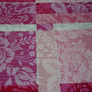 May include: Close-up of a fabric with a floral pattern in shades of pink, red, and white. The design features large rose and leaf motifs, arranged in a patchwork style with stripes and solid blocks.