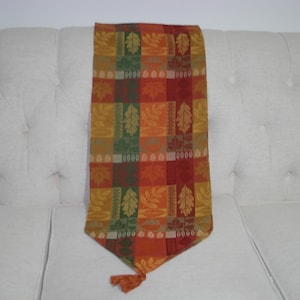 May include: A rectangular table runner featuring a patchwork design of autumn-themed squares in shades of orange, red, yellow, and green. The fabric displays repeating patterns of maple and oak leaves, with a pointed end finished with a tassel.