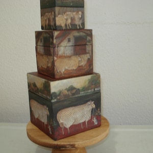 May include: A stack of three square wooden boxes featuring hand-painted farm scenes. The bottom box displays a large sheep, the middle box shows a rural landscape with a barn, and the top box depicts a group of sheep in a field with muted tones.