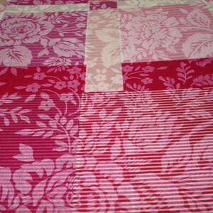 May include: A patchwork quilt with floral designs in pink, red, and white. The quilt features large roses, small flowers, and striped details. The design combines various textures and patterns, creating a visually appealing textile.