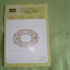May include: A white card with a gold decorative design in the shape of a wreath. The card is labeled "Big Shot" and has the text "Shown at 50% Mostrado al 50% Echelle: 50% Abbildung in 50% der Originalgröße".