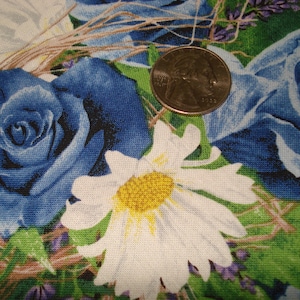 May include: Close-up of a floral fabric featuring large blue roses, white daisies with yellow centres, and green leaves. A U.S. quarter coin rests on the fabric, providing a size reference. The background includes lavender-coloured accents.