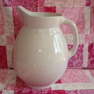 May include: A white ceramic pitcher with a rounded body and a handle. The pitcher has a smooth, glossy finish.