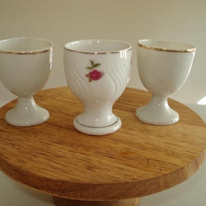 May include: Three white porcelain egg cups with gold trim, displayed on a round wooden stand. One cup features a pink floral design. The egg cups are a classic design.