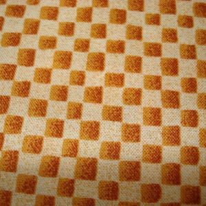 May include: Close-up of a fabric with a repeating checkered pattern. The squares alternate between a light beige and a warm, burnt orange color. The fabric has a textured appearance, suggesting a woven material.