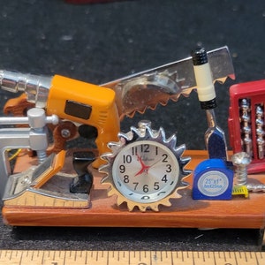 May include: A wooden base with a clock, a drill, a saw, a ruler, a clamp, and a toolbox with drill bits. The clock face reads "11 12 1 2 3 4 5 6 7 8 9 10". The ruler is marked "MADE IN U.S.A. 1 2 H P 3 4". The toolbox is red and has a silver handle. The drill is orange and black. The saw is silver and has a black handle. The clamp is silver and has a black handle. The ruler is wood and has black markings.