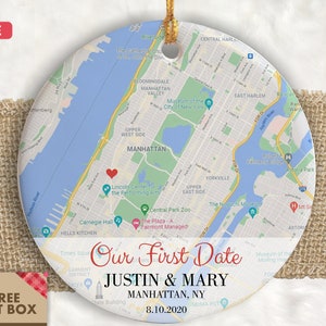 May include: A round, white ceramic ornament with a map of Manhattan, New York City. The map shows the location of Central Park, the Lincoln Center for the Performing Arts, and Carnegie Hall. The ornament is decorated with the text "Our First Date" and the names "Justin & Mary" along with the date "8.10.2020".