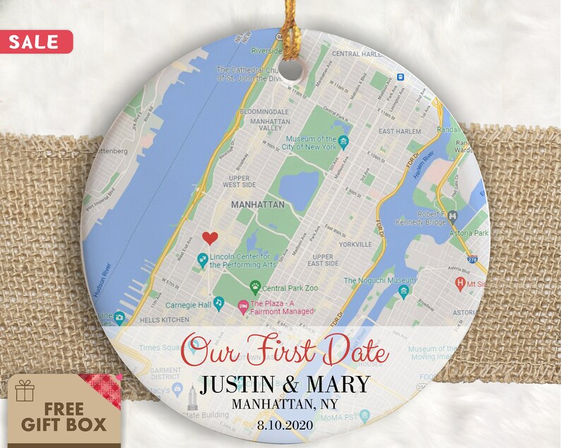 May include: A round, white ceramic ornament with a map of Manhattan, New York City. The map shows the location of Central Park, the Lincoln Center for the Performing Arts, and Carnegie Hall. The ornament is decorated with the text "Our First Date" and the names "Justin & Mary" along with the date "8.10.2020".