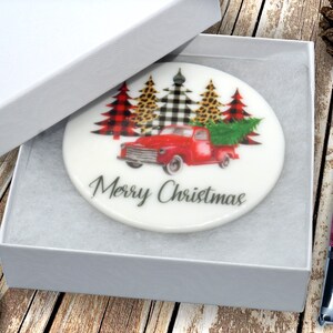 May include: White ceramic ornament with a red truck carrying a Christmas tree. The truck is surrounded by three trees decorated with buffalo plaid, black and white checkered, and leopard print. The ornament reads "Merry Christmas".