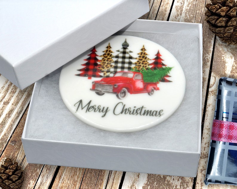May include: White ceramic ornament with a red truck carrying a Christmas tree. The truck is surrounded by three trees decorated with buffalo plaid, black and white checkered, and leopard print. The ornament reads "Merry Christmas".