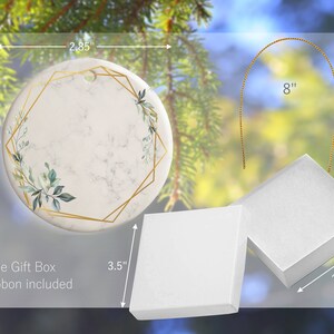 May include: A white ceramic ornament with a gold geometric frame and green foliage design. The ornament is 2.85 inches in diameter and comes with a 8 inch gold ribbon and a white gift box.
