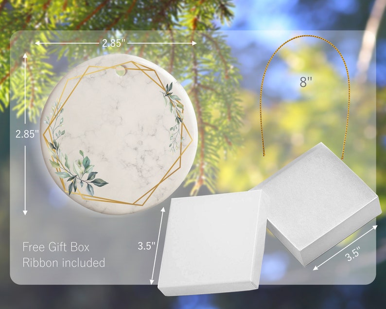 May include: A white ceramic ornament with a gold geometric frame and green foliage design. The ornament is 2.85 inches in diameter and comes with a 8 inch gold ribbon and a white gift box.