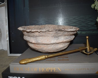 May include: A textured, light brown decorative bowl with a layered design. A gold-colored, decorative object with a handle and a pointed end rests on a book. The book's title is "HOUSE OF HOPPEN".