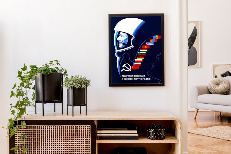 Może przedstawiać: Winietkowy plakat propagandowy Związku Radzieckiego przedstawiający astronautę w hełmie na niebieskim tle. Plakat zawiera tekst "Мы дружим и создаем, и в космос мир утверждаем!" kt&oacute;ry tłumaczy się jako "Jesteśmy przyjaci&oacute;łmi i tworzymy, i potwierdzamy pok&oacute;j w kosmosie!" Plakat zawiera r&oacute;wnież szereg flag reprezentujących r&oacute;żne kraje.