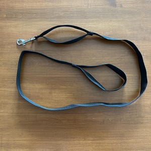 May include: A black dog lead with a silver metal clasp. The lead is made of a woven material and has a loop handle. The lead is lying on a wooden surface.