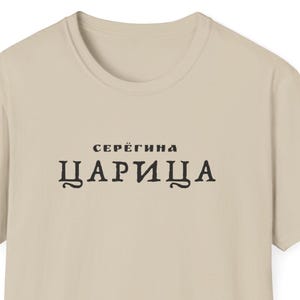 Könnte beinhalten: Sandfarbenes T-Shirt mit Rundhalsausschnitt. Auf der Vorderseite des Shirts steht der schwarze Text "СЕРЁГИНА ЦАРИЦА". Das Wort "SAND" befindet sich oben links. Das Shirt besteht aus einem weichen Material.