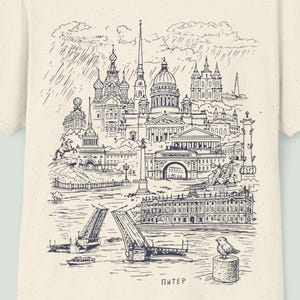 May include: Cream-colored t-shirt featuring a detailed black and white illustration of a cityscape, including buildings, bridges, and water. The word "Питер" is printed at the bottom. The shirt has short sleeves and a crew neck.