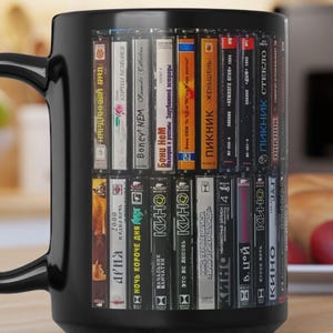 Può includere: Tazza da caffè nera da 15 once con un design di una libreria piena di videocassette. Le cassette hanno vari titoli in russo. La tazza ha un manico grande ed è su una superficie di legno.