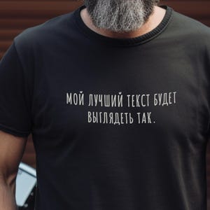 Op de afbeelding: Zwart T-shirt met ronde hals en witte tekst: "МОЙ ЛУЧШИЙ ТЕКСТ БУДЕТ ВЫГЛЯДЕТЬ ТАК." Het T-shirt wordt gedragen door een persoon met een grijze baard.