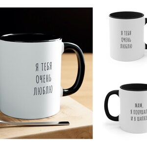 May include: Two white ceramic mugs with black rims. The mug on the left has the text "Я ТЕБЯ ОЧЕНЬ ЛЮБЛЮ" printed in black. The mug on the right has the text "MAM. Я ПОКУШАЛ И В ШАПКЕ" printed in black.