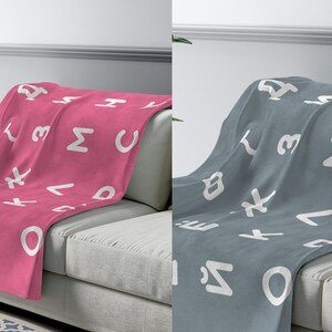 May include: Two fleece blankets with a white alphabet pattern on a pink and gray background. The blankets are draped over a light gray couch.