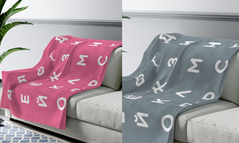 May include: Two fleece blankets with a white alphabet pattern on a pink and gray background. The blankets are draped over a light gray couch.