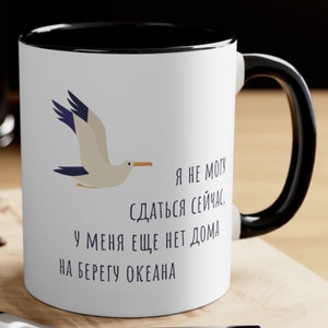 Può includere: Una tazza in ceramica bianca con manico e bordo neri. La tazza presenta un gabbiano disegnato in volo e il testo "Я не могу сдаться сейчас. У меня еще нет дома на берегу океана."
