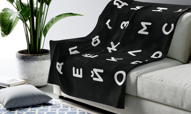 May include: A black fleece throw blanket with a white alphabet pattern. The blanket is draped over a white couch.