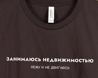 以下が含まれることがあります： 白い文字で「Занимаюсь недвижимостью лежу и не двигаюсь」と書かれた濃い茶色のTシャツです。