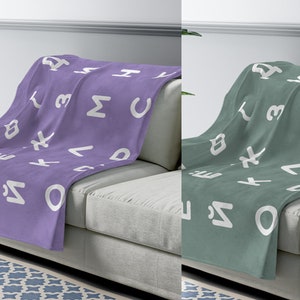 May include: Two fleece blankets with a white alphabet pattern on a light gray couch. One blanket is purple and the other is green.