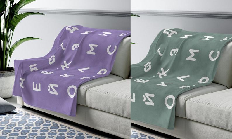 May include: Two fleece blankets with a white alphabet pattern on a light gray couch. One blanket is purple and the other is green.