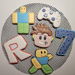 May include: A collection of six decorated sugar cookies featuring two block-style figures with yellow heads, a white game controller with blue buttons, a letter R with a pink outline, a character head with brown hair, and a blue number 7.