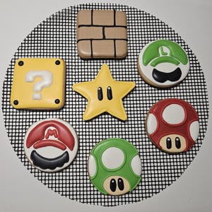 May include: A collection of seven decorated sugar biscuits featuring iconic video game imagery, including a yellow question block, a tan brick block, a yellow star, a red mushroom, a green mushroom, and two circular character hats in red and green.