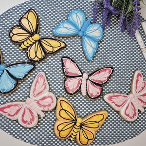May include: Six butterfly-shaped sugar biscuits decorated with royal icing in yellow, light blue, and pink, featuring black outlines and detailed wing patterns. Two white edible ink markers with yellow and black accents are positioned to the right.