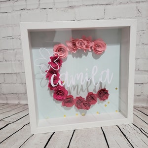 May include: A white shadow box with a light blue background. The box contains a paper flower arrangement in shades of pink, forming the letter "C" with the name "Camila" written in white below the flowers. Gold glitter is scattered on the bottom of the box.