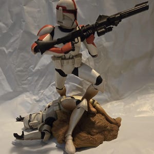 May include: A Star Wars action figure set featuring a standing clone trooper with orange and white armor holding a long black blaster rifle, positioned over a fallen clone trooper on a brown rocky base, all rendered in a detailed plastic finish.