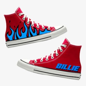 以下が含まれることがあります： 赤いハイカットスニーカー。片方の靴には青い炎が描かれ、もう片方の靴には青い文字で「BILLIE」と書かれている