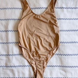 May include: A tan-colored one-piece swimsuit with a scoop neckline and thin straps. The swimsuit is made of a smooth, shiny fabric and has a high-cut leg opening. The garment is laid flat on a white and blue striped fabric.