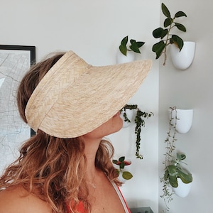 Op de afbeelding: Een vrouw draagt een lichtbruine strooien zonneklep met een brede rand. De zonneklep is gemaakt van geweven stro en heeft een eenvoudig ontwerp.